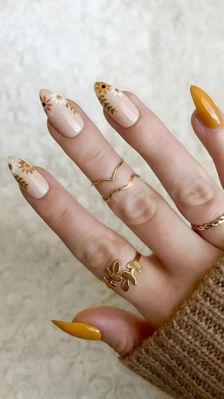 Hand with spring floral press-on nails in nude and mustard yellow, wearing gold rings